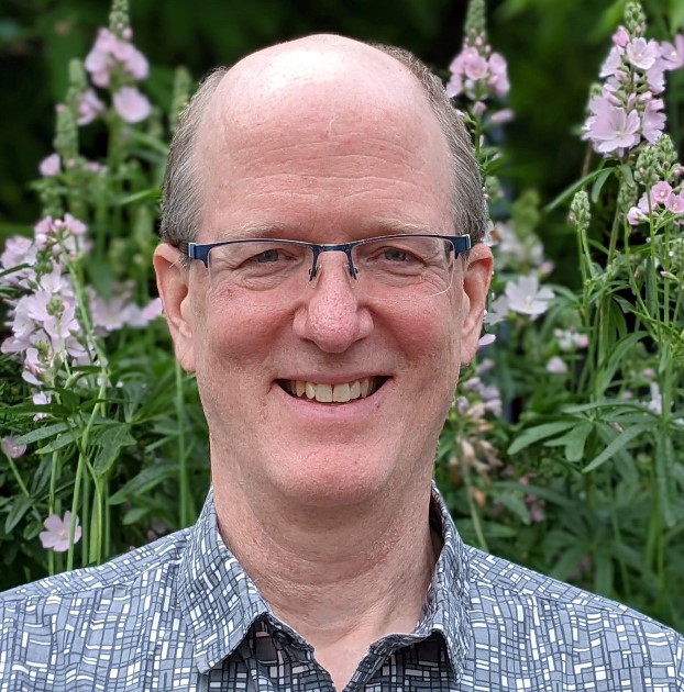a white man in glasses smiles into the camera. He's surrounded by pink flowers in the background.